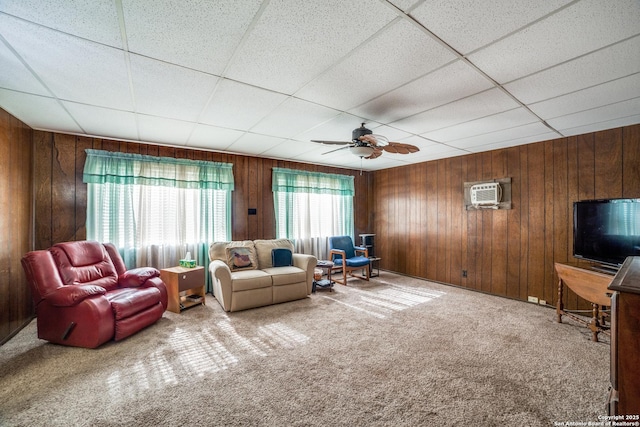 living room featuring wooden walls, a drop ceiling, a ceiling fan, a wall unit AC, and carpet floors