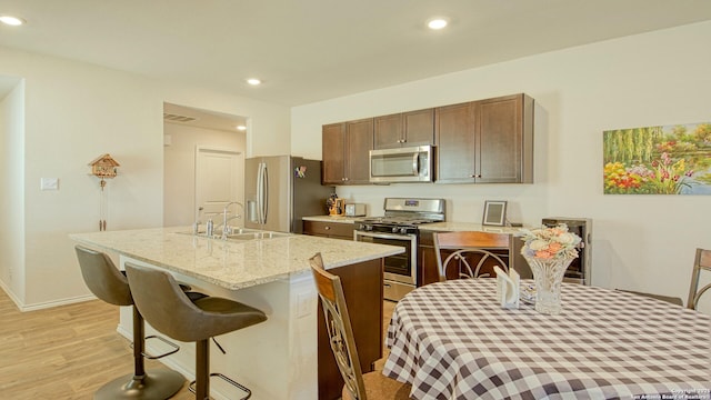 kitchen with appliances with stainless steel finishes, sink, light stone countertops, a center island with sink, and light wood-type flooring