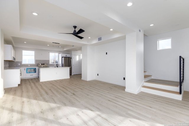 unfurnished living room with a raised ceiling, ceiling fan, sink, and light wood-type flooring