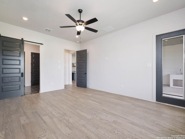 unfurnished bedroom with ensuite bath, a barn door, ceiling fan, and light wood-type flooring