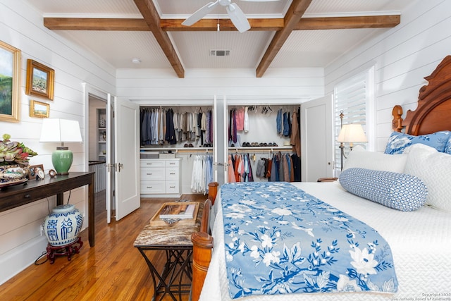 bedroom featuring light hardwood / wood-style flooring, ceiling fan, wooden walls, two closets, and beamed ceiling