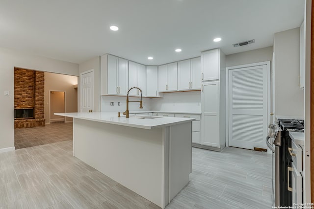 kitchen with stainless steel gas stove, sink, white cabinets, a kitchen island with sink, and a brick fireplace