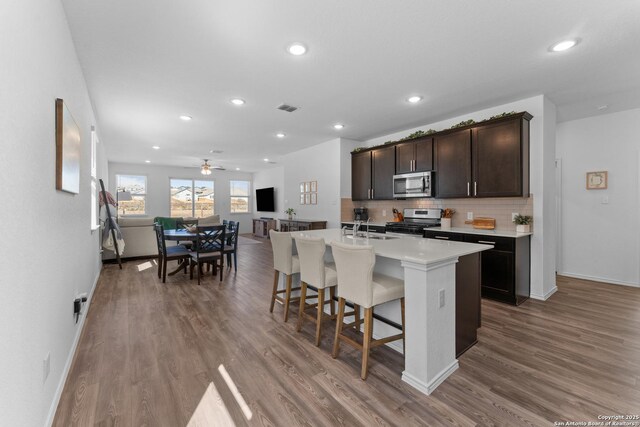 kitchen with appliances with stainless steel finishes, a breakfast bar, wood-type flooring, backsplash, and a kitchen island with sink