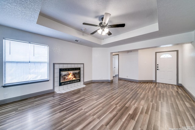 unfurnished living room featuring wood-type flooring, a tiled fireplace, ceiling fan, a tray ceiling, and a textured ceiling