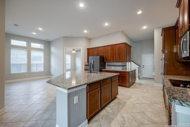 kitchen with stainless steel appliances, light stone countertops, a kitchen island with sink, and ceiling fan