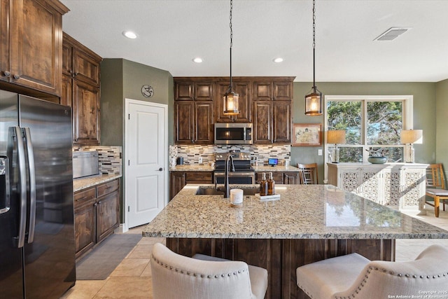 kitchen with light stone counters, stainless steel appliances, decorative light fixtures, and an island with sink