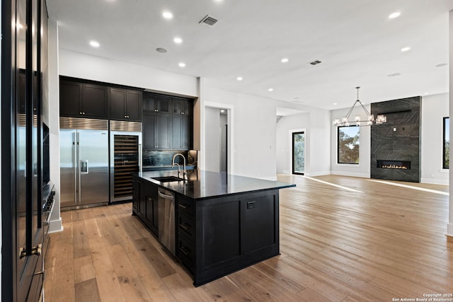 kitchen featuring sink, appliances with stainless steel finishes, an island with sink, beverage cooler, and light wood-type flooring