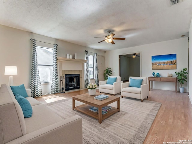 living room with a tile fireplace, ceiling fan, a textured ceiling, and light hardwood / wood-style flooring