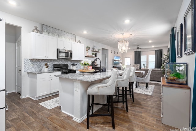 kitchen with hanging light fixtures, light stone counters, an island with sink, white cabinets, and black range with gas stovetop