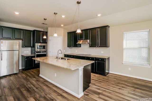 kitchen with sink, light stone counters, hanging light fixtures, appliances with stainless steel finishes, and a kitchen island with sink