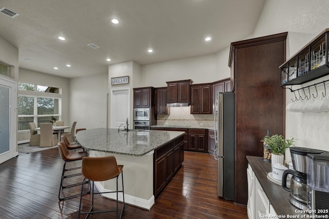 kitchen featuring backsplash, dark hardwood / wood-style flooring, stainless steel appliances, light stone countertops, and a center island with sink