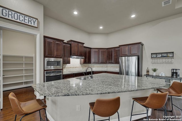 kitchen featuring appliances with stainless steel finishes, dark hardwood / wood-style flooring, a kitchen island with sink, light stone counters, and dark brown cabinets