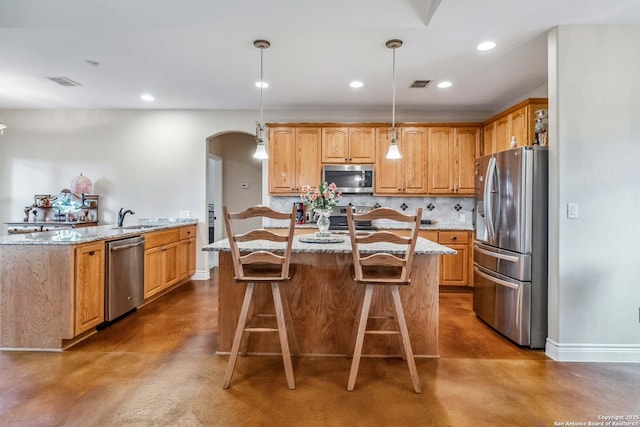 kitchen featuring appliances with stainless steel finishes, hanging light fixtures, light stone countertops, an island with sink, and decorative backsplash