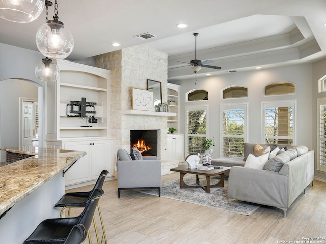 living room featuring a stone fireplace, built in features, ornamental molding, ceiling fan, and light hardwood / wood-style floors