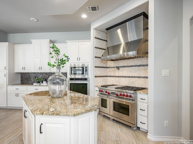 kitchen with appliances with stainless steel finishes, white cabinetry, light stone countertops, a kitchen island, and wall chimney exhaust hood