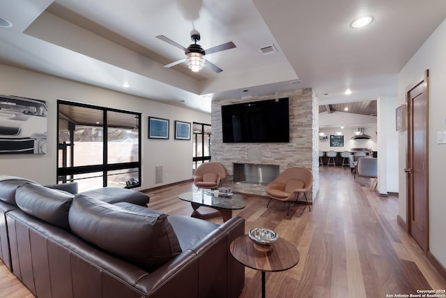 living room featuring a tray ceiling, a fireplace, ceiling fan, and light wood-type flooring