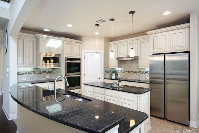 kitchen featuring appliances with stainless steel finishes, decorative light fixtures, a kitchen island with sink, and dark stone countertops