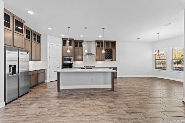 kitchen with tasteful backsplash, decorative light fixtures, a center island with sink, stainless steel appliances, and wall chimney range hood