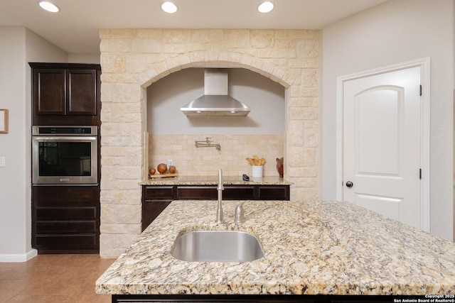 kitchen featuring wall chimney exhaust hood, sink, stainless steel oven, an island with sink, and light stone countertops