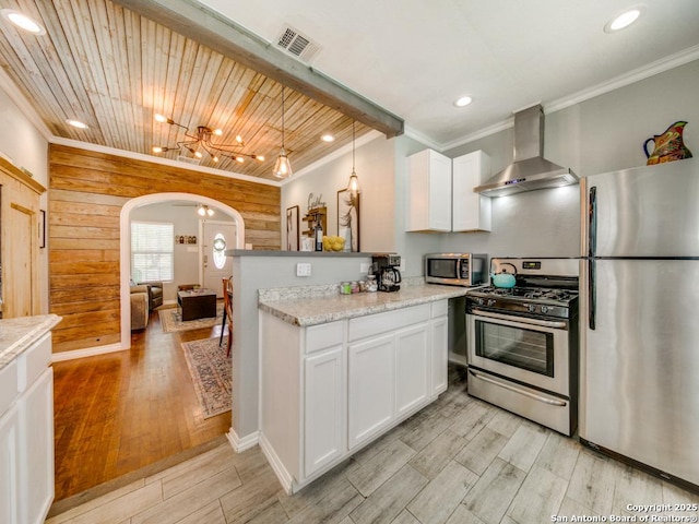 kitchen featuring wall chimney range hood, appliances with stainless steel finishes, white cabinetry, ornamental molding, and kitchen peninsula