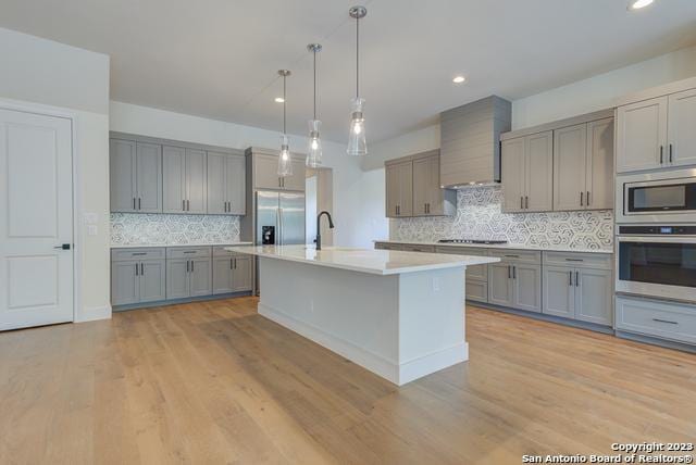 kitchen featuring gray cabinets, pendant lighting, an island with sink, built in appliances, and wall chimney range hood