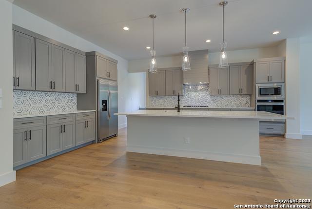 kitchen featuring gray cabinets, hanging light fixtures, a kitchen island with sink, stainless steel appliances, and wall chimney exhaust hood