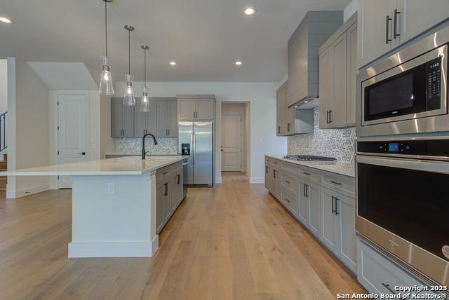 kitchen featuring appliances with stainless steel finishes, gray cabinets, an island with sink, pendant lighting, and light hardwood / wood-style floors