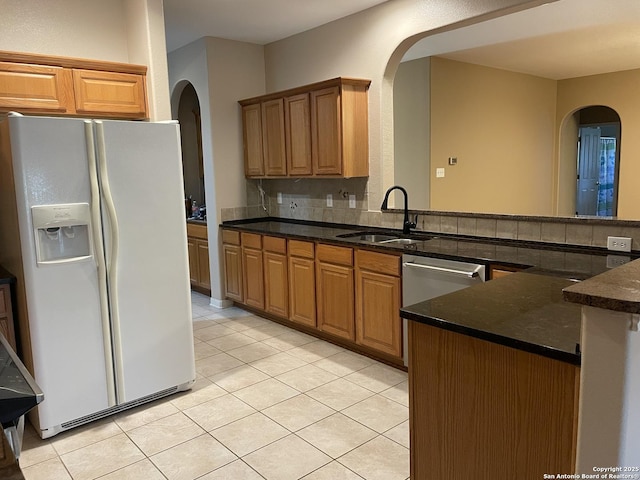 kitchen with sink, white refrigerator with ice dispenser, light tile patterned flooring, stainless steel dishwasher, and dark stone counters