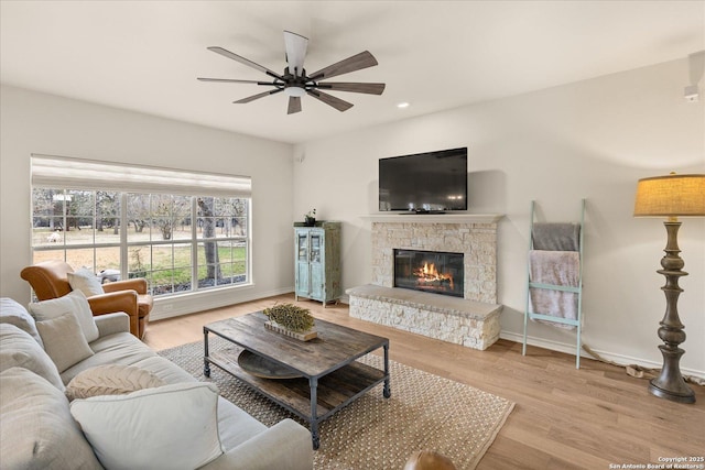 living room featuring a ceiling fan, baseboards, wood finished floors, and a stone fireplace