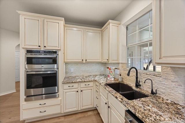 kitchen featuring arched walkways, backsplash, cream cabinets, double oven, and a sink