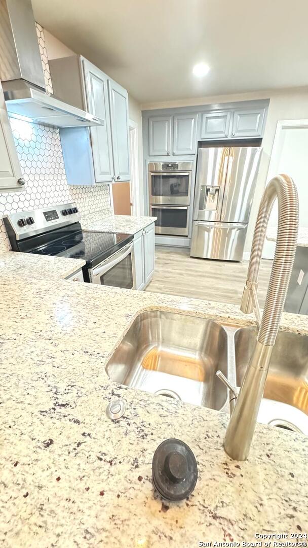 kitchen featuring wall chimney exhaust hood, sink, light stone counters, stainless steel appliances, and backsplash