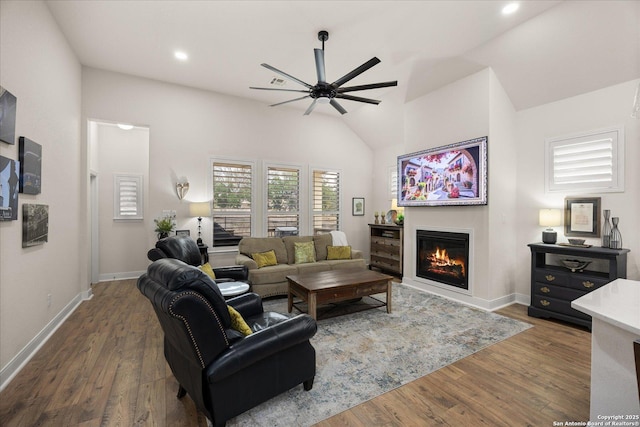 living area featuring a ceiling fan, a glass covered fireplace, wood finished floors, high vaulted ceiling, and baseboards