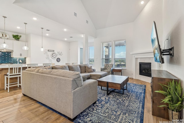 living area featuring high vaulted ceiling, light wood-style flooring, visible vents, and a glass covered fireplace