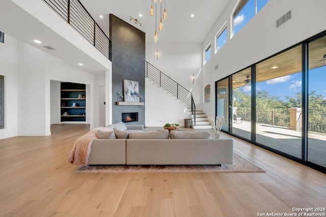 living room featuring built in features, a towering ceiling, a notable chandelier, a large fireplace, and light wood-type flooring