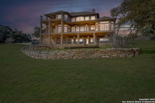 back house at dusk featuring a balcony, ceiling fan, and a lawn