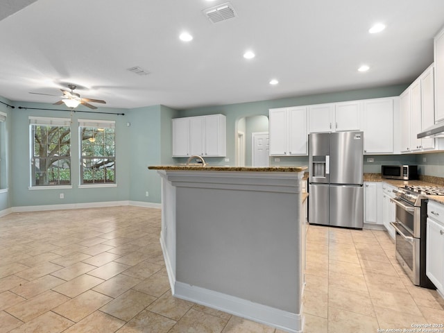 kitchen featuring ceiling fan, appliances with stainless steel finishes, dark stone countertops, an island with sink, and white cabinets