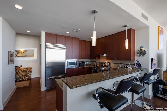 kitchen featuring decorative light fixtures, dark stone countertops, built in appliances, kitchen peninsula, and dark wood-type flooring