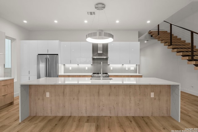 kitchen featuring pendant lighting, wall chimney range hood, high end fridge, white cabinets, and a large island with sink