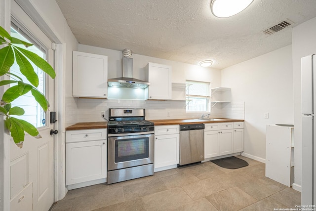 kitchen with wall chimney range hood, wooden counters, appliances with stainless steel finishes, white cabinetry, and backsplash