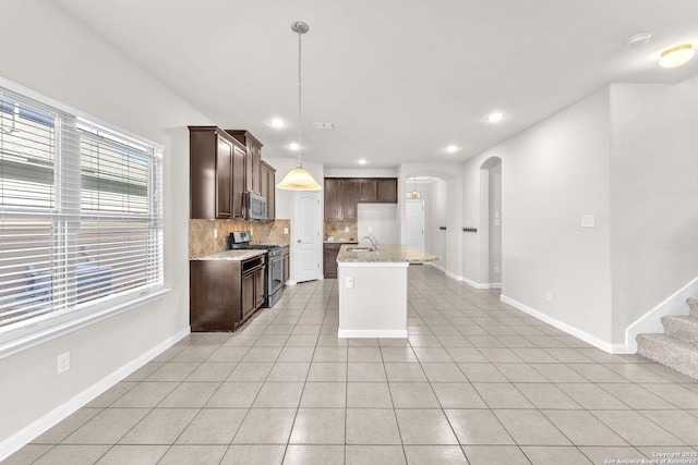 kitchen with decorative light fixtures, a kitchen island with sink, light tile patterned floors, stainless steel appliances, and light stone countertops