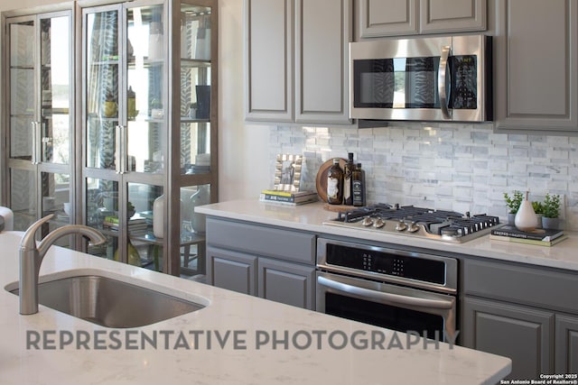 kitchen featuring sink, gray cabinetry, backsplash, stainless steel appliances, and light stone countertops