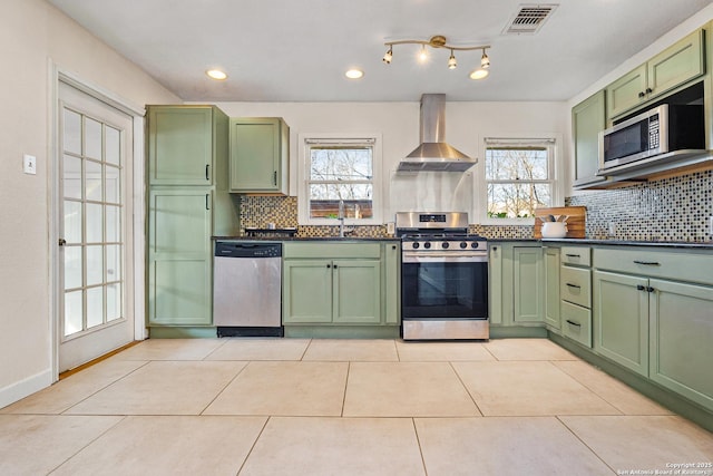 kitchen featuring light tile patterned flooring, wall chimney exhaust hood, green cabinets, stainless steel appliances, and backsplash