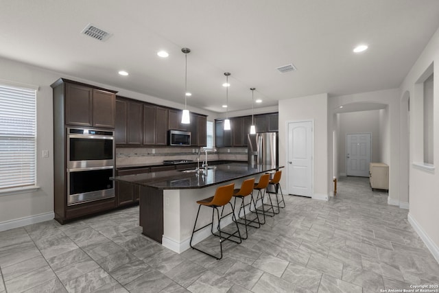 kitchen with a kitchen island with sink, hanging light fixtures, backsplash, dark stone countertops, and stainless steel appliances