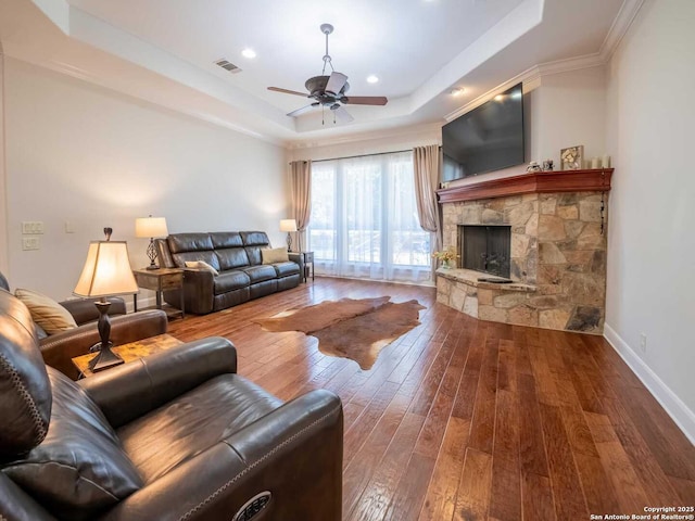 living room with crown molding, ceiling fan, a tray ceiling, wood-type flooring, and a stone fireplace