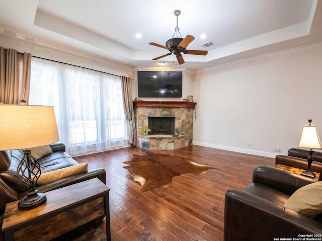 living room with ceiling fan, a tray ceiling, a stone fireplace, and dark wood-type flooring