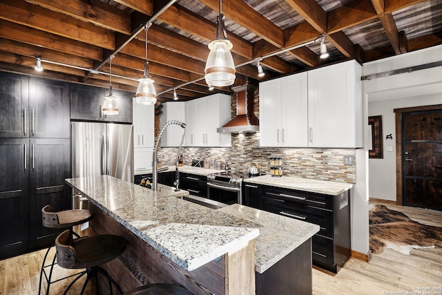 kitchen featuring appliances with stainless steel finishes, tasteful backsplash, an island with sink, hanging light fixtures, and wall chimney exhaust hood