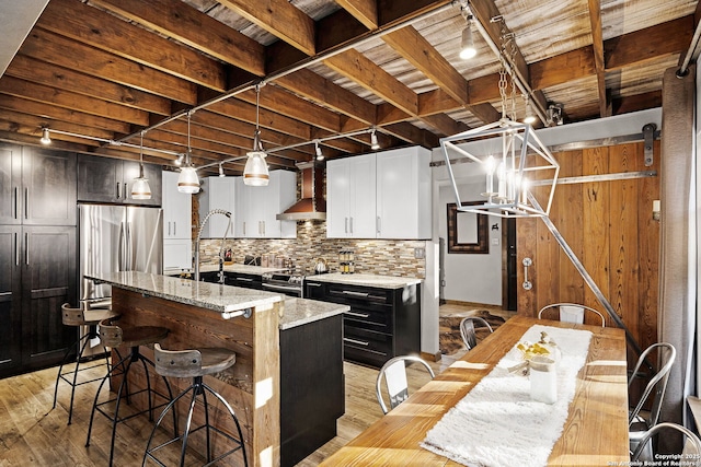 kitchen with appliances with stainless steel finishes, pendant lighting, a center island with sink, light wood-type flooring, and wall chimney exhaust hood