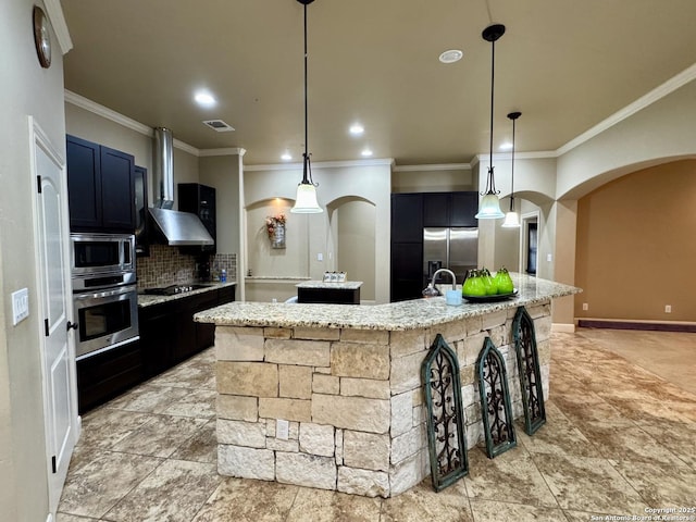 kitchen with pendant lighting, appliances with stainless steel finishes, an island with sink, and wall chimney range hood