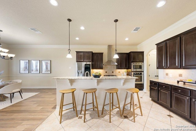 kitchen with an island with sink, a breakfast bar, appliances with stainless steel finishes, wall chimney range hood, and pendant lighting