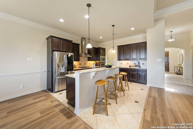 kitchen featuring appliances with stainless steel finishes, light stone countertops, wall chimney range hood, decorative light fixtures, and a kitchen island with sink